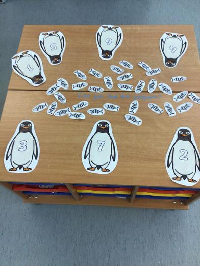 Counting activity with paper penguins showing numbers and many fish cut-outs scattered on a wooden table. 