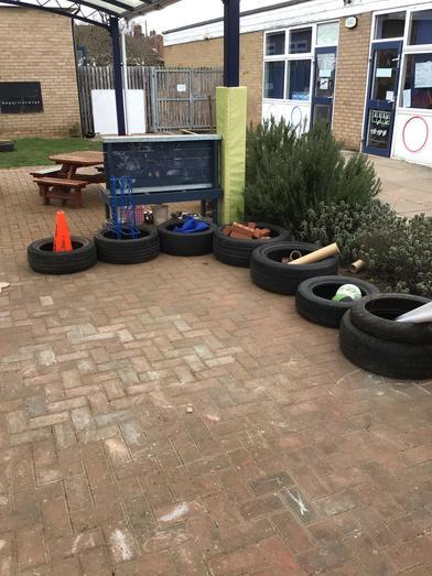 Outdoor school playground with brick patio, many recycled tyres, picnic bench, art station, and brick building with blue doors. 