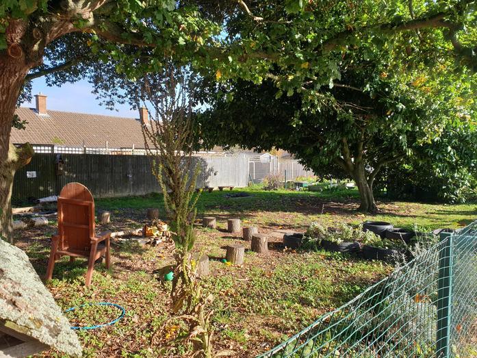 A natural outdoor garden or play area with large trees, grassy ground, wooden tree stumps, a wooden chair, and a green fence. 