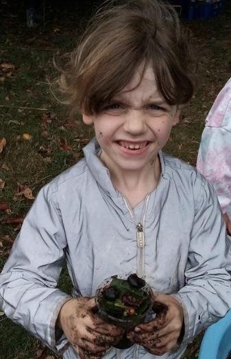 Close-up of a smiling, mud-covered child in a grey jacket, holding a container of leaves and mud outdoors. 