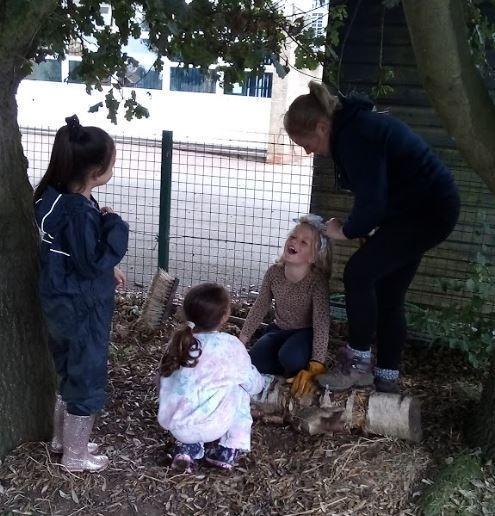 An adult brushes the hair of a laughing young girl sitting on a log outdoors. Two other children watch nearby in a natural play area. 