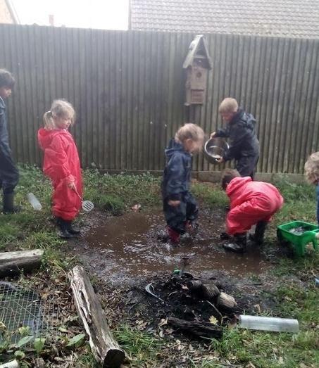 Young children in waterproofs happily splash and play in a large muddy puddle outdoors, surrounded by grass and a fence. 
