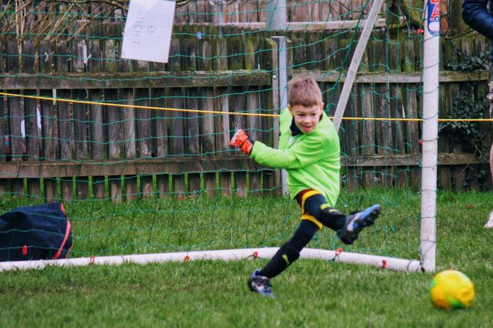 A young boy goalkeeper in a green shirt and orange gloves kicks a yellow football in a garden, smiling, with a goal net behind him. 