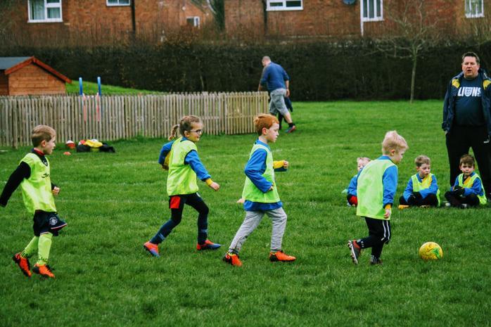 Young children in bright bibs play football on a green field. An adult coach stands nearby. 
