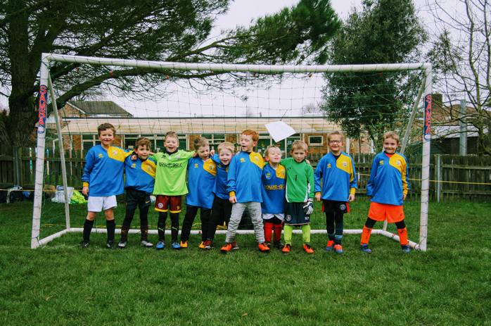 A cheerful children's football team, wearing blue, yellow, and green kits, stands smiling in front of a football goal on a grassy pitch. 