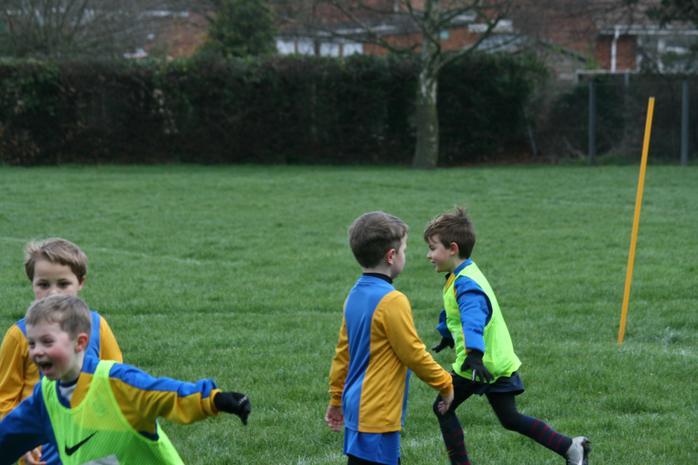 Excited young boys in football kits and bibs playing on a grassy field with a training pole nearby. 